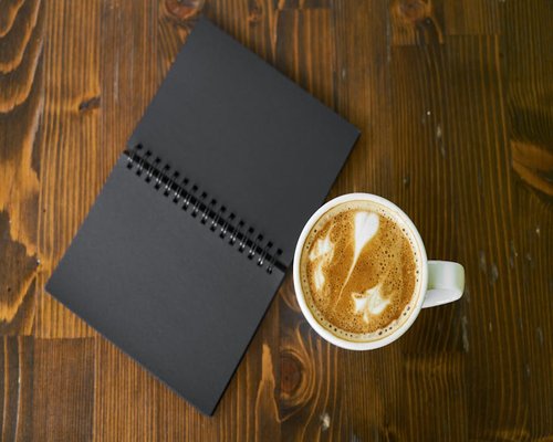 A freshly brewed warm beverage sits on a wooden workspace table next to a notebook